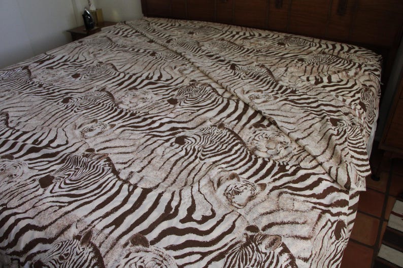 Puede incluir: Una s&aacute;bana con un patr&oacute;n repetitivo de rayas de cebra en marr&oacute;n y blanco. La tela muestra m&uacute;ltiples cabezas de cebra integradas en el dise&ntilde;o a rayas, cubriendo toda la superficie de la ropa de cama extendida sobre una estructura de cama grande.