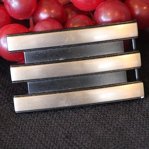 May include: A silver and black belt buckle with a rectangular design. The buckle has three horizontal silver bars and a black base. The background features red grapes on a dark surface.