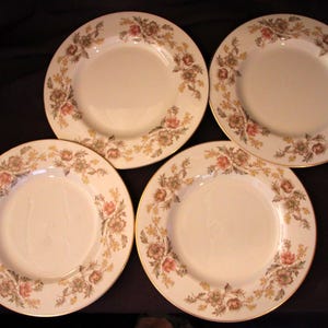 May include: A set of four round ceramic plates featuring a cream-colored center and a decorative border of muted pink, brown, and yellow floral patterns. Each plate is finished with a thin gold-toned rim along the outer edge of the circular design.