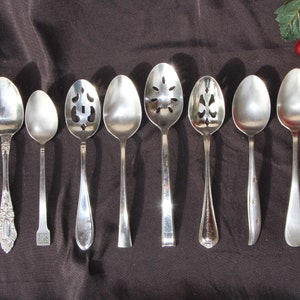 May include: Eight silver spoons with different designs, including slotted spoons and a spoon with a decorative handle. The spoons are arranged in a row on a dark brown surface.