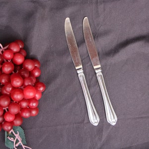May include: Two silver dinner knives with ornate handles on a dark gray tablecloth next to a bunch of red grapes.