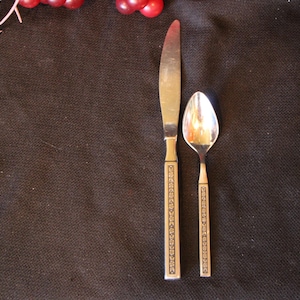 May include: A stainless steel knife and spoon set with decorative handles. The handles have a gold-colored design. The silverware is placed on a dark brown surface, with a few red berries in the upper left corner.