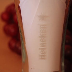 May include: A clear glass beer vessel featuring the Heineken brand name and a star logo etched into the surface. The glass has a tapered cylindrical shape with a smooth texture and transparent finish, displaying the brand text vertically.
