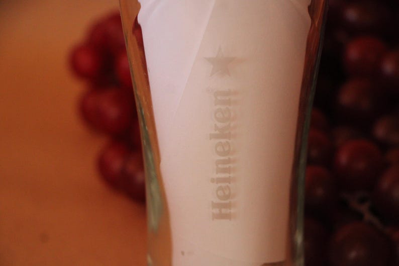 May include: A clear glass beer vessel featuring the Heineken brand name and a star logo etched into the surface. The glass has a tapered cylindrical shape with a smooth texture and transparent finish, displaying the brand text vertically.