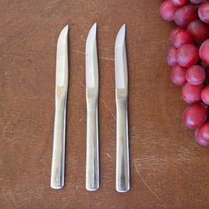 May include: Three silver butter knives on a brown wooden surface.