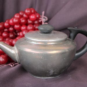 May include: A silver teapot with a black handle and a lid. The teapot is sitting on a purple cloth with red berries in the background.