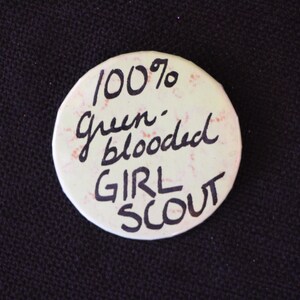 May include: A white button with the text "100% Green-blooded Girl Scout" written in black ink.