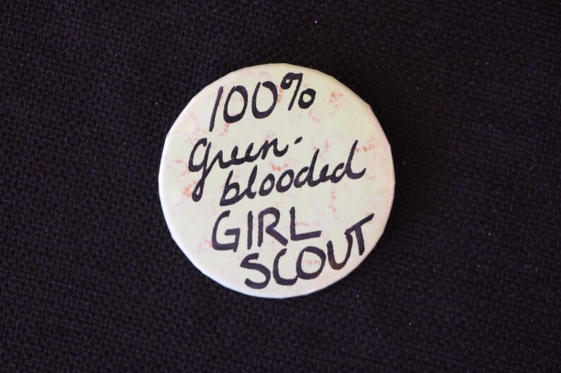 May include: A white button with the text "100% Green-blooded Girl Scout" written in black ink.