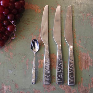 May include: A set of stainless steel silverware, including three knives and a spoon, with textured handles. A bunch of red grapes is in the upper left corner. The silverware is arranged on a distressed green surface.