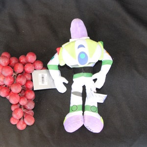 May include: A plush toy of Buzz Lightyear from the Toy Story franchise. The toy is green, white, and purple with a black belt. The toy is standing on a black surface.