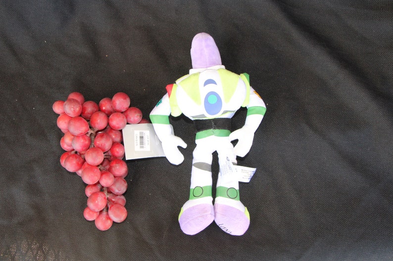 May include: A plush toy of Buzz Lightyear from the Toy Story franchise. The toy is green, white, and purple with a black belt. The toy is standing on a black surface.
