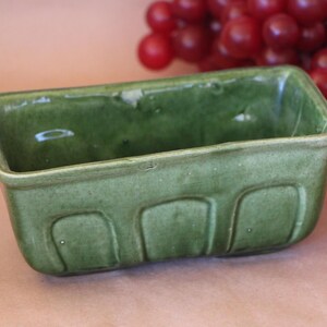 May include: A rectangular ceramic loaf pan with a glossy olive green glaze. The exterior features a series of three molded, recessed arch panels along the front side. The interior is smooth and deep, with a consistent green finish throughout the piece.