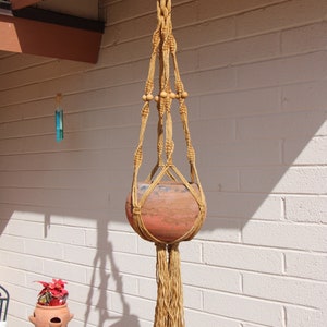 May include: A hanging macrame plant holder with a terracotta pot. The macrame is made of tan rope and features wooden beads. The pot is reddish-brown and suspended from a metal ring attached to a brown overhang.