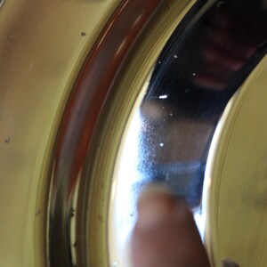 Puede incluir: Primer plano de una secci&oacute;n de un plato o cuenco de color dorado, posiblemente de lat&oacute;n. La superficie es reflectante, con una banda roja oscura y un reflejo oscuro. La imagen est&aacute; bien iluminada, mostrando los detalles del metal.