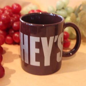 May include: A dark brown mug with the word "HEY'S" in large, white capital letters. The mug is surrounded by red and green grapes. The mug has a handle and is likely used for drinking beverages.
