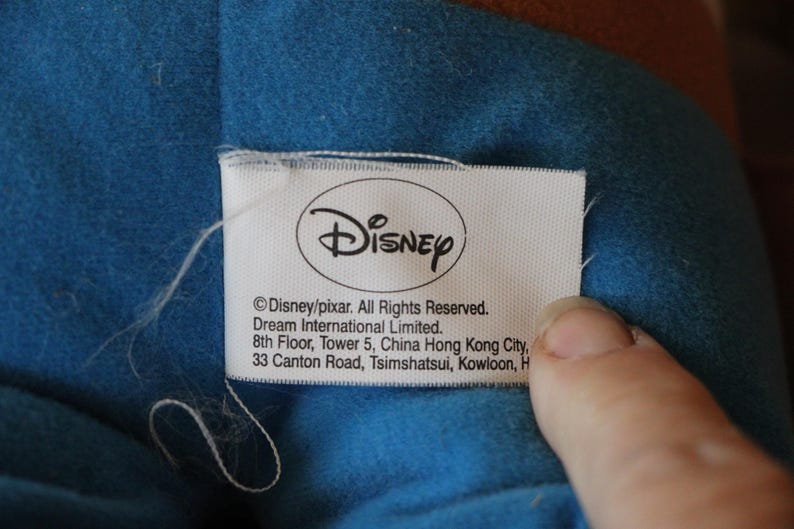 Puede incluir: Una etiqueta blanca con el logotipo de Disney y el texto "Disney/pixar. Todos los derechos reservados. Dream International Limited. 8&ordm; piso, Torre 5, China Hong Kong City, 33 Canton Road, Tsimshatsui, Kowloon, H"