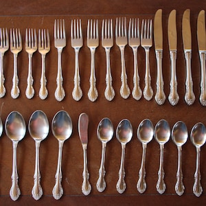 May include: A set of silverware, including forks, spoons, and knives, laid out on a wooden surface. The silverware is made of polished metal and has an ornate design.