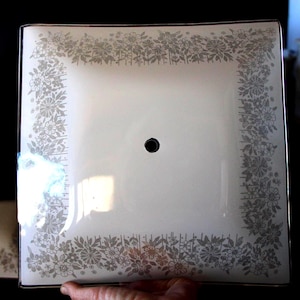 May include: Square, white glass light fixture with a floral pattern border in shades of gray. The fixture has a black center hole and a black border. The floral design is repeated around the edges of the square.