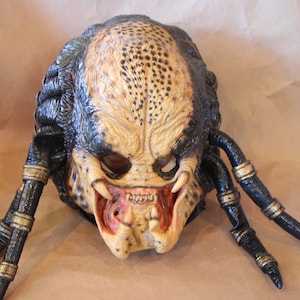 May include: A detailed Predator mask with a textured, tan and black design. The mask features prominent mandibles, tusks, and dreadlocks. The mask is a prop from the Predator movie franchise.