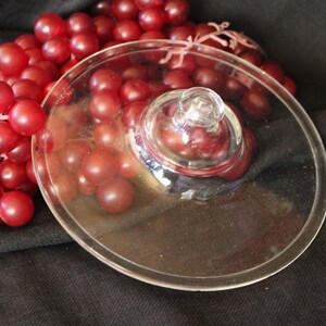 May include: A clear glass lid with a knob sits atop a cluster of red grapes. The grapes are a deep red color and are partially covered by the glass lid. The background is a dark fabric.