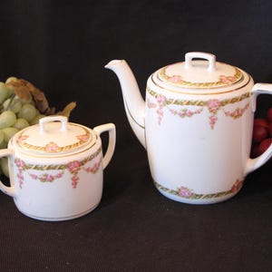 May include: A white porcelain teapot and creamer set with a floral design. The teapot has a gold trim and a pink and green floral pattern. The creamer has a matching design.