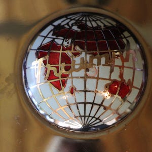 Puede incluir: Un objeto decorativo dorado con un dise&ntilde;o de globo terr&aacute;queo. El globo tiene un fondo blanco con un patr&oacute;n de cuadr&iacute;cula y continentes rojos. La palabra "Wilson" se muestra en letras doradas en el globo.