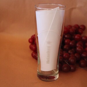May include: A clear glass beer tumbler featuring the Heineken logo and a star printed in white on the front. A piece of white paper is inserted inside the glass, and a cluster of dark red grapes is positioned directly behind the glass.