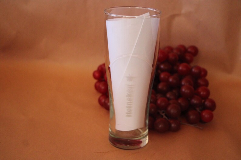 May include: A clear glass beer tumbler featuring the Heineken logo and a star printed in white on the front. A piece of white paper is inserted inside the glass, and a cluster of dark red grapes is positioned directly behind the glass.