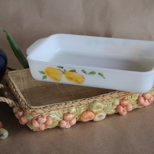 May include: White rectangular baking dish with a floral design featuring yellow lemons and green leaves. The dish is sitting on a woven basket with a peach-colored floral design.