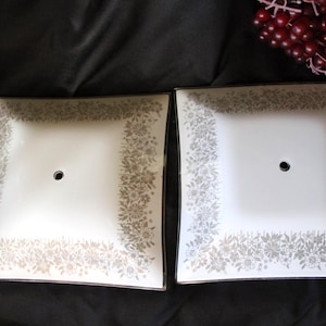 May include: Two square, vintage ceiling light covers. Each has a white center with a black hole and a silver floral design border. The covers are on a dark background.