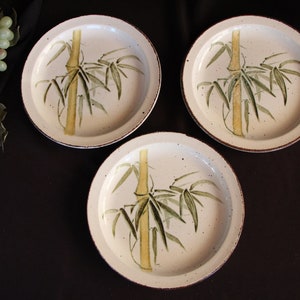 May include: Three white ceramic dinner plates with a brown speckled glaze. Each plate features a hand-painted design of a bamboo stalk with green leaves.