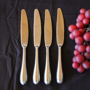 May include: Four silver butter knives with rounded handles are arranged in a row on a black surface. A bunch of red grapes is to the right of the knives.