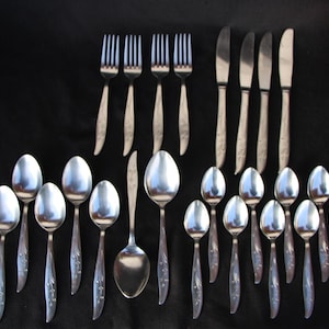 May include: A collection of vintage silverware, including forks, knives, and spoons, arranged on a black surface. The silverware has a floral design on the handles and a shiny, reflective surface. A cluster of red grapes is in the upper right corner.