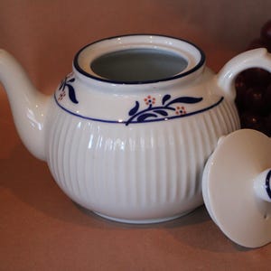 May include: A white ceramic teapot with a ribbed body, a curved spout, and a handle. The teapot features a blue horizontal line near the top rim, accented with small blue leaf patterns and tiny orange dots. A matching white lid with a blue knob is included.