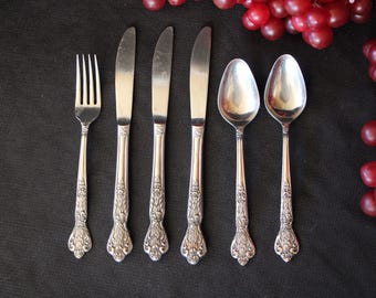 May include: A set of vintage silverware, including a fork, knives, and spoons, arranged on a dark surface. The silverware has ornate handles and is accompanied by a cluster of red grapes, creating an elegant dining scene.