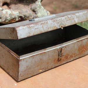 May include: A vintage metal toolbox with a hinged lid, showing signs of rust and wear. The toolbox is open and empty, revealing the interior.