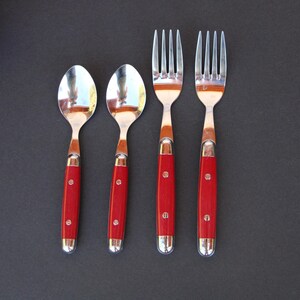 May include: Two silver-toned metal spoons and two silver-toned metal forks arranged side by side. Each utensil features a bright red handle secured with two small metal rivets. The spoons have rounded bowls and the forks have four tines.