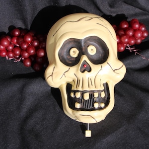 May include: A cream-colored skull-shaped wall decoration with black eye sockets and mouth. A small red accent is in the nose. Two bunches of red grapes are on either side of the skull against a black background.