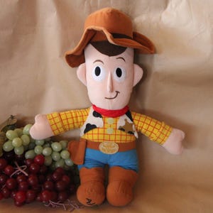 Puede incluir: Un juguete de peluche de Woody de Toy Story. Lleva su caracter&iacute;stico sombrero de vaquero marr&oacute;n, camisa amarilla y azul y botas marrones. Tiene una sonrisa amigable y sostiene una bandana roja.