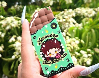 May include: A rectangular chocolate bar-shaped accessory with a brown textured top and a light green wrapper. The wrapper features a cartoon character with dark hair and horns, the text Awooooo and Milk Chocolate, and small paw print icons.