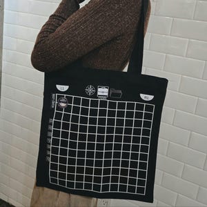 Puede incluir: Un bolso de tela negro con un patrón de cuadrícula blanca que recuerda a una pantalla de inventario de videojuego. El diseño incluye iconos de una brújula, un maletín etiquetado como ITEMS, una carpeta, botones L1 y R1, y un gráfico de cinta.