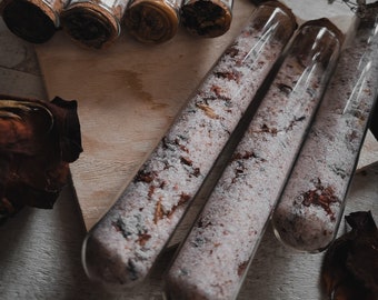 May include: Six glass test tubes filled with pink bath salts and dried rose petals. The test tubes have cork stoppers and are arranged on a wooden surface.