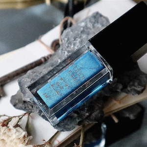 May include: A clear rectangular makeup container with a black cap, containing blue eyeshadow with the text "Maiden Earth". The container rests on a gray stone, with a vintage key and dried flowers nearby.