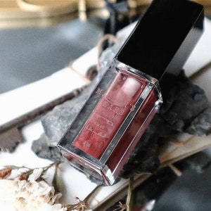 May include: A rectangular lipstick tube with a black cap and a clear body, filled with a deep red liquid. The tube is labeled with the text "MADARS". The lipstick is resting on a white surface with a vintage key and dried flowers.