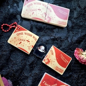 May include: Rectangular acrylic keychains with red and cream designs featuring the text ONE WISH WILLOW, CRACK, AMAZE YOUR FRIENDS, and You only get ONE WISH. One keychain is broken in two, revealing a small black character charm in the centre.