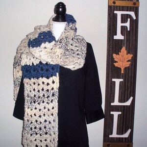 May include: A crocheted scarf in shades of cream, blue, and gray. The scarf is draped over a black mannequin and has a textured, chunky stitch pattern.