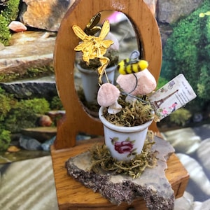 May include: A miniature wooden vanity with a small drawer, a mirror, and a white ceramic flower pot with pink mushrooms and a yellow bee. The vanity is decorated with moss and a gold fairy figurine.