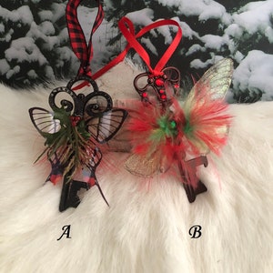 May include: Two Christmas ornaments shaped like keys with butterfly wings. The key on the left has black wings with green and red accents. The key on the right has red, green, and gold wings. Both keys have red ribbon.