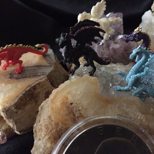 May include: A collection of small, colorful dragon figurines, including red, black, and blue, are displayed on a bed of light-colored rocks and crystals. The dragons have gold accents and are posed in various positions. A small, pink gem is visible.