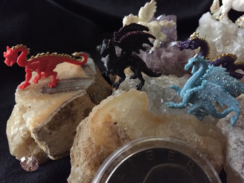 May include: A collection of small, colorful dragon figurines, including red, black, and blue, are displayed on a bed of light-colored rocks and crystals. The dragons have gold accents and are posed in various positions. A small, pink gem is visible.